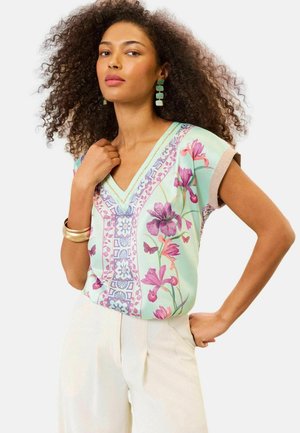 Kobieta z kręconymi włosami, ubraną w zieloną kwiecistą bluzkę z dekoltem w serek, szerokie kremowe spodnie, zielone kolczyki i złote bransoletki, pozująca z ręką na biodrze.