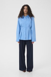 Kobieta o kręconych włosach, ubrana w jasnoniebieską zapinaną na guziki koszulę i ciemne, szerokie spodnie, stojąca na jednolitym białym tle.