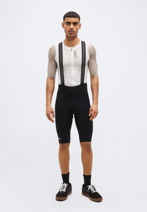 Muž stojaci, oblečený v bielom cyklistickom drese s béžovými rukávmi a čiernych šortkách s trakmi, obutý v čiernych teniskách, na jednoduchom svetlom pozadí.