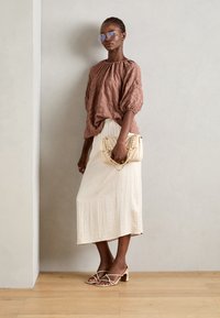 Braune strukturierte Bluse, cremefarbene Rock, beige Handtasche mit Kette und Riemchenabsätze mit kleinem Blockabsatz. Transparente Sonnenbrille als Akzent.
