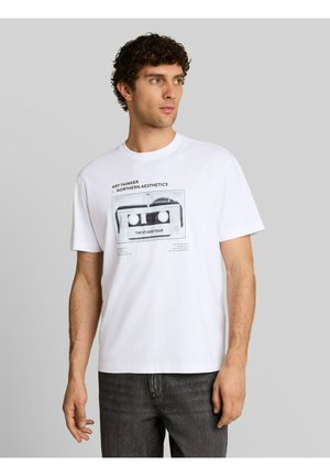 Mann trägt ein weißes T-Shirt mit Kassettenspieler-Grafik und Text "Art Thinker Northern Aesthetics", blickt nach rechts mit neutralem Gesichtsausdruck.