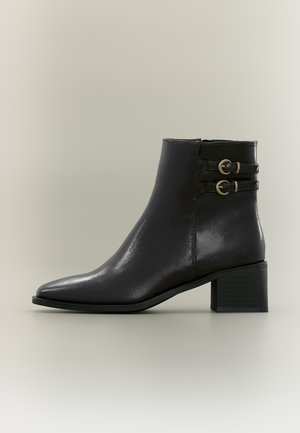 Bottine en cuir noir avec petit talon bloc et deux sangles décoratives avec boucles dorées sur le côté.