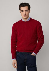 Pull en maille rouge avec un col rond et un ourlet côtelé. Porté par-dessus une chemise à col bleu clair, associé à un jean foncé.