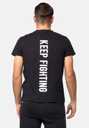 Homme aux cheveux courts et bruns portant un t-shirt noir avec un texte vertical blanc "KEEP FIGHTING" dans le dos, debout devant un fond uni.