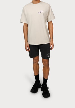 Jasno beżowa koszulka bawełniana z purpurowym logo, w połączeniu z czarnymi szortami z graficznym nadrukiem, czarnymi skarpetkami i masywnymi czarnymi sneakersami.