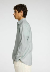 Camicia a maniche lunghe con bottoni in tessuto verde chiaro, presenta un colletto a punta, una vestibilità rilassata e una sottile texture. Indossata con pantaloni color crema.