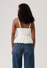 Haut peplum blanc avec un tissu texturé et des bretelles fines, mettant en valeur un corps smocké. Associé à un jean en denim décontracté de couleur foncée.