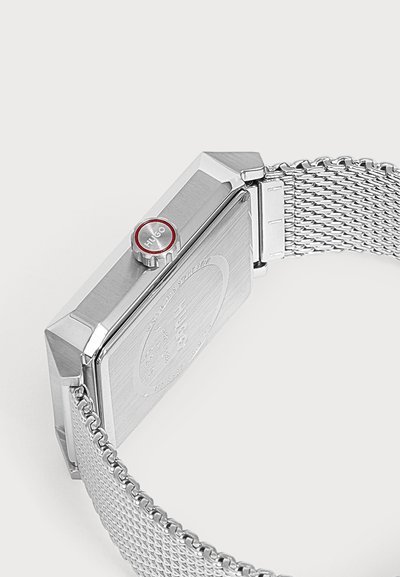 Montre à bracelet en maille métallique argentée avec boîtier rectangulaire et couronne accentuée de rouge gravée avec "Hugo" sur fond uni.