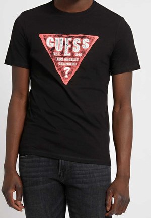Persona che indossa una maglietta nera con un logo triangolare rosso di Guess e testo bianco con la scritta "Guess Est 1981 Los Angeles California?"