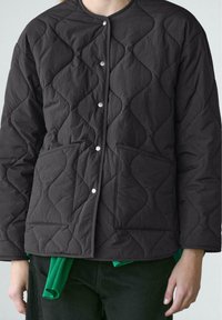 Quiltet sort jakke med skjulte trykknapper foran og to påsyede lommer, båret over en grøn sweater og sorte bukser.