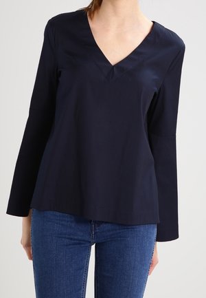 Kvinde iført en mørkeblå langærmet V-hals bluse kombineret med blå denimjeans, stående mod en ensfarvet baggrund.