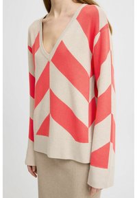 Pull à col en V beige et corail avec de grandes rayures en chevron, coupe ample, bords côtelés et manches longues. Texture en tricot douce.