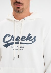 Sudadera blanca con capucha, que presenta un logo en azul oscuro de "Creeks" y el texto "Original Desde 1974", con cordones y una textura suave.