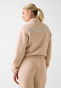Beige cropped sweatshirt met "GUESS" in glanzende strass steentjes op de rug, met een hoge kraag en elastische manchetten.