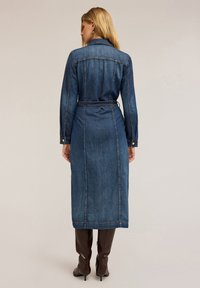 Donkerblauwe denim jurk met lange mouwen, een ceintuur op de taille en een ontwerp tot op de grond. Voorzien van zilveren knopen en een zachte structuur.