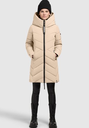Beige puffer coat with chevron stitching, high collar, and front zip closure. Paired with black leggings and knee-high black boots.