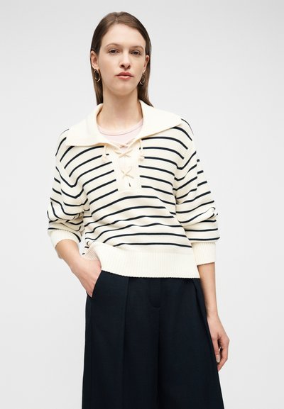 Γυναίκα με πουλόβερ σε κρέμ και μαύρες ρίγες με δέσιμο στο γιακά και σκούρο παντελόνι με φαρδιά μπατζάκια, στέκεται με το ένα χέρι στην τσέπη.