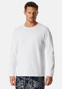 Camiseta blanca de manga larga con cuello redondo y un logo sutil en el lado izquierdo del pecho, con una textura de tela suave y lisa.