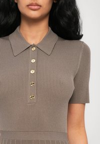 Gros plan sur une femme portant un polo côtelé taupe à manches courtes, avec un col et cinq boutons dorés sur le devant.