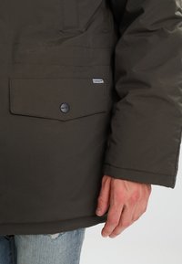 Giacca imbottita verde scuro con tasca a pattina dotata di un bottone nero e un piccolo etichetta con logo. Il tessuto appare testurizzato e durevole.