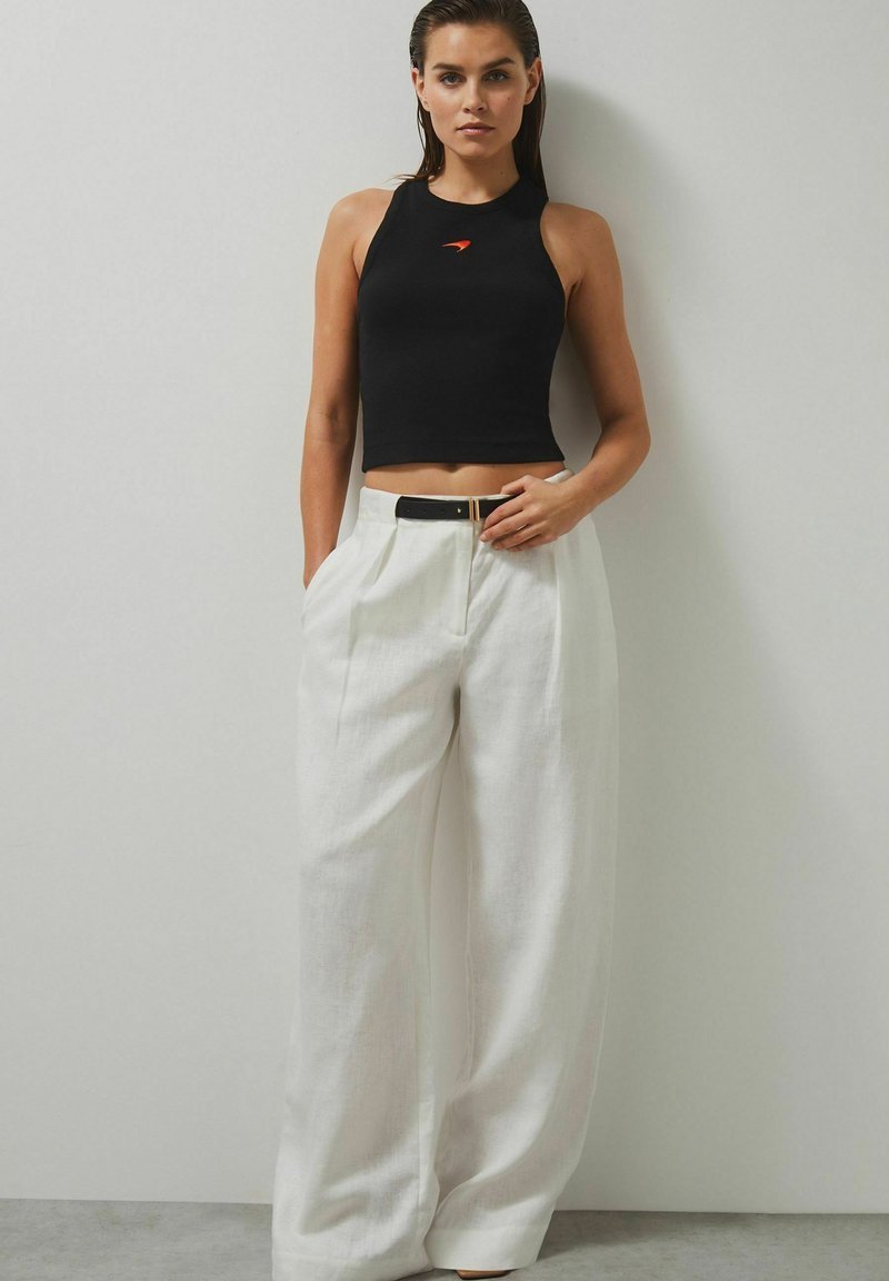 Svart ärmlös crop-top med en framsida logotyp och hög halsringning, kombinerad med vita vida byxor och ett svart bälte.