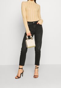 Maglione beige a coste con collo alto, jeans neri a vita alta e tacchi neri con cinturini. Tiene una piccola borsa metallica con dettagli scintillanti.