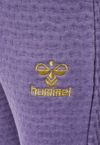 Paarse textuurstof met een gouden geborduurd logo met een gestileerd ontwerp boven het woord "hummel" linksonder.
