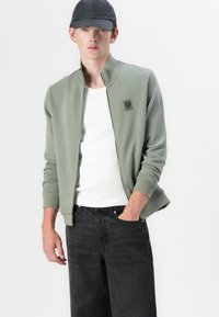 Light green zip-up jacket, ribbed cuffs, and collar, featuring a logo patch. Worn over a white tank top and paired with dark jeans.