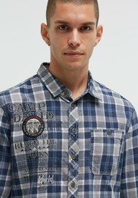 Blau- und graues Karo-Hemd mit Knopfleiste und Kragen, mit gesticktem Text und einem Patch auf der Brusttasche. Glattes Baumwollgewebe.