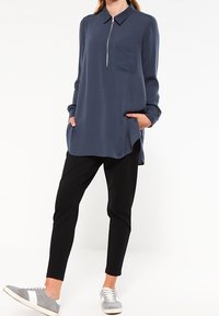 Blouse à manches longues bleu marine avec un col et des détails de fermeture éclair, associée à un pantalon noir slim et des baskets grises. Dispose d'une poche poitrine.