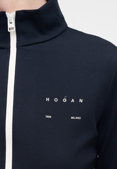Σκούρο μπλε μπουφάν με φερμουάρ και λευκή επένδυση στο φερμουάρ και λευκή επιγραφή "HOGAN 1986 MILANO" στο αριστερό στήθος.