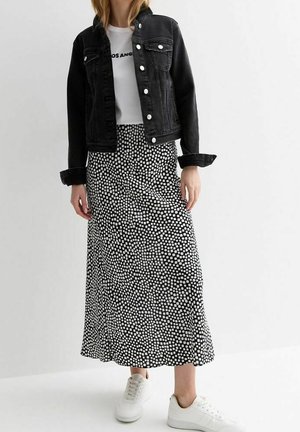 Femme portant une veste en jean noire, un t-shirt blanc graphique, une jupe midi noire à pois blancs, et des baskets blanches, debout devant un fond uni.