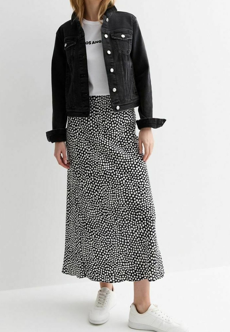 Femme portant une veste en jean noire, un t-shirt blanc graphique, une jupe midi noire à pois blancs, et des baskets blanches, debout devant un fond uni.