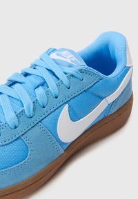 Sinine sametist spordijalats, millel on võrgusilma aktsendid, valge Nike logo, lame pael ja pruun kummitald. Omab tugevdusõmblusi.