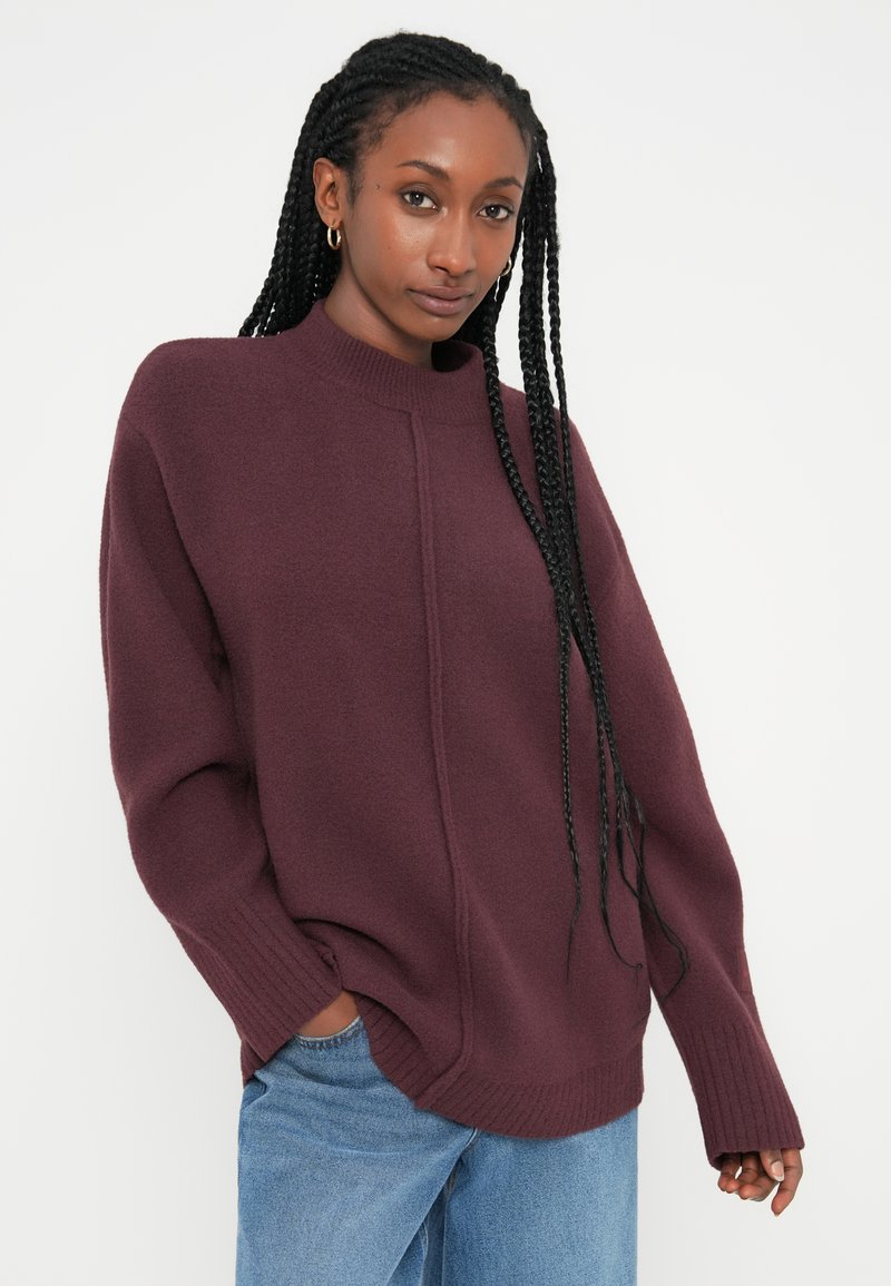 Burgundy stickad tröja med hög krage, ribbade ärmavslut och sidofickor, tillsammans med ljusblå jeans. Mjuk textur och avslappnad passform.