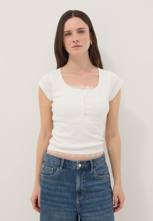 Giovane donna con lunghi capelli castani indossa una maglietta bianca a maniche corte con bordatura in pizzo e jeans blu a vita alta, in piedi davanti a uno sfondo semplice.