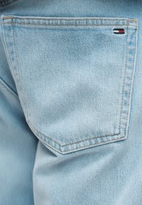 Πίσω τσέπη ανοιχτού μπλε τζιν με μικρό ορθογώνιο λογότυπο της μάρκας Tommy Hilfiger σε κόκκινο, άσπρο και μπλε στην επάνω δεξιά γωνία.