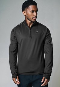 Kwartrits lange mouw top in donkergrijze stof, met een subtiele logo op de borst en effen grijze contrasterende accenten op de schouders.