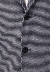 Ναυτικό μπλε υφασμάτινο σακάκι με πλεκτό σχέδιο, διαθέτοντας δύο μαύρους κουμπιά και λεπτομέρεια τσέπης. Κατασκευασμένο από ένα ελαφρύ ύφασμα.