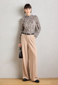 Langärmliges Oberteil mit einem beige-schwarzen Wellenmuster, kombiniert mit beigen weit geschnittenen Hosen, einem schwarzen Gürtel und einer kleinen schwarzen Handtasche.