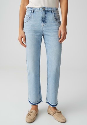 Vrouw draagt lichtblauwe jeans met rechte pijpen, rafelige zomen en beige loafers, gecropt onder de taille.