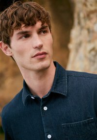 Donker denim overhemd met knoopsluiting, voorzien van een kraag, een borstzak en witte knopen. De stof heeft een gladde textuur met subtiele vervagingdetails.