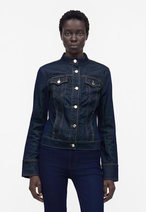 Femeie purtând o jachetă din denim albastru închis, strâmtă, cu nasturi argintii și pantaloni din denim asortați, stând în fața unui fundal alb.