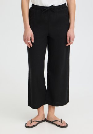 Person trägt lockere schwarze kurze Hosen mit Kordelzug in der Taille, weißes Oberteil, Ringe an den Fingern und schwarze Flip-Flops vor weißem Hintergrund.