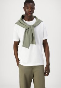 Witte t-shirt met korte mouwen, gecombineerd met olijfgroene broek, met een bijpassende groene sjaal om de schouders gedrapeerd. Glad stofoppervlak.