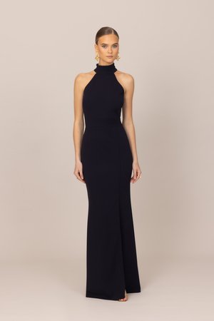 Femme portant une robe de soirée noire sans manches à col montant, avec des boucles d'oreilles dorées imposantes, debout devant un fond beige uni.