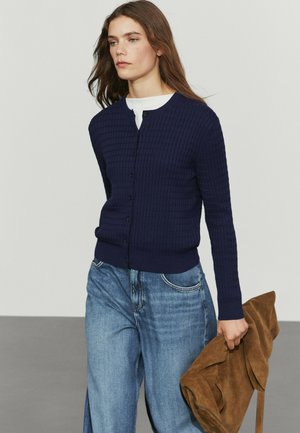 Donna che indossa un cardigan blu navy con bottoni, jeans azzurri, che tiene una grande borsa marrone in camoscio su uno sfondo chiaro uniforme.