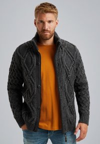 Cardigan gris de punto trenzado con ribete negro, que presenta un cuello alto, cierres de botón y puños acanalados, superpuesto a una camiseta de manga larga mostaza.