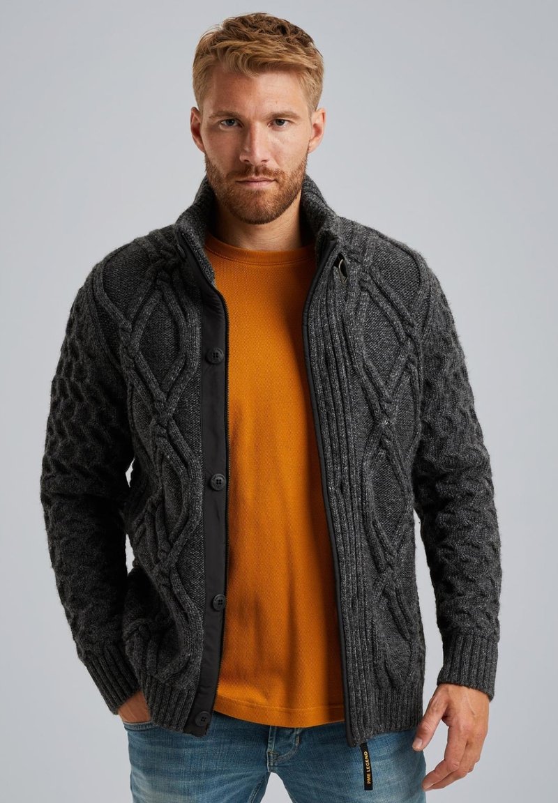 Cardigan gris de punto trenzado con ribete negro, que presenta un cuello alto, cierres de botón y puños acanalados, superpuesto a una camiseta de manga larga mostaza.