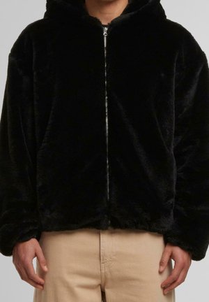 Personne portant un sweat à capuche noir en tissu pelucheux avec fermeture éclair et un pantalon beige, debout avec les bras détendus le long du corps.